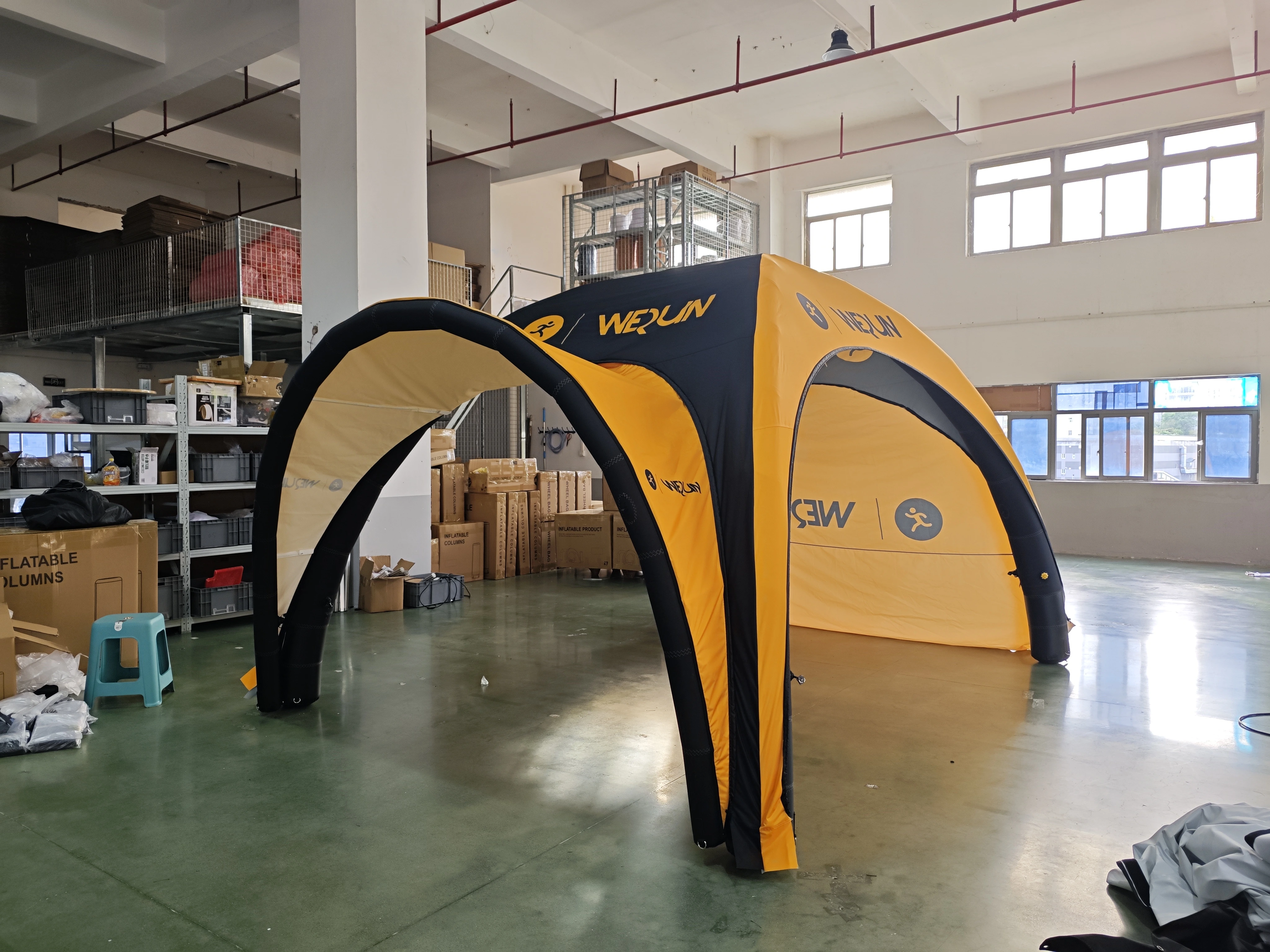 An indoor perspective of a yellow and black "WEQUN" branded inflatable arch tent, shown set up inside a spacious industrial factory or warehouse with large windows and high ceilings.